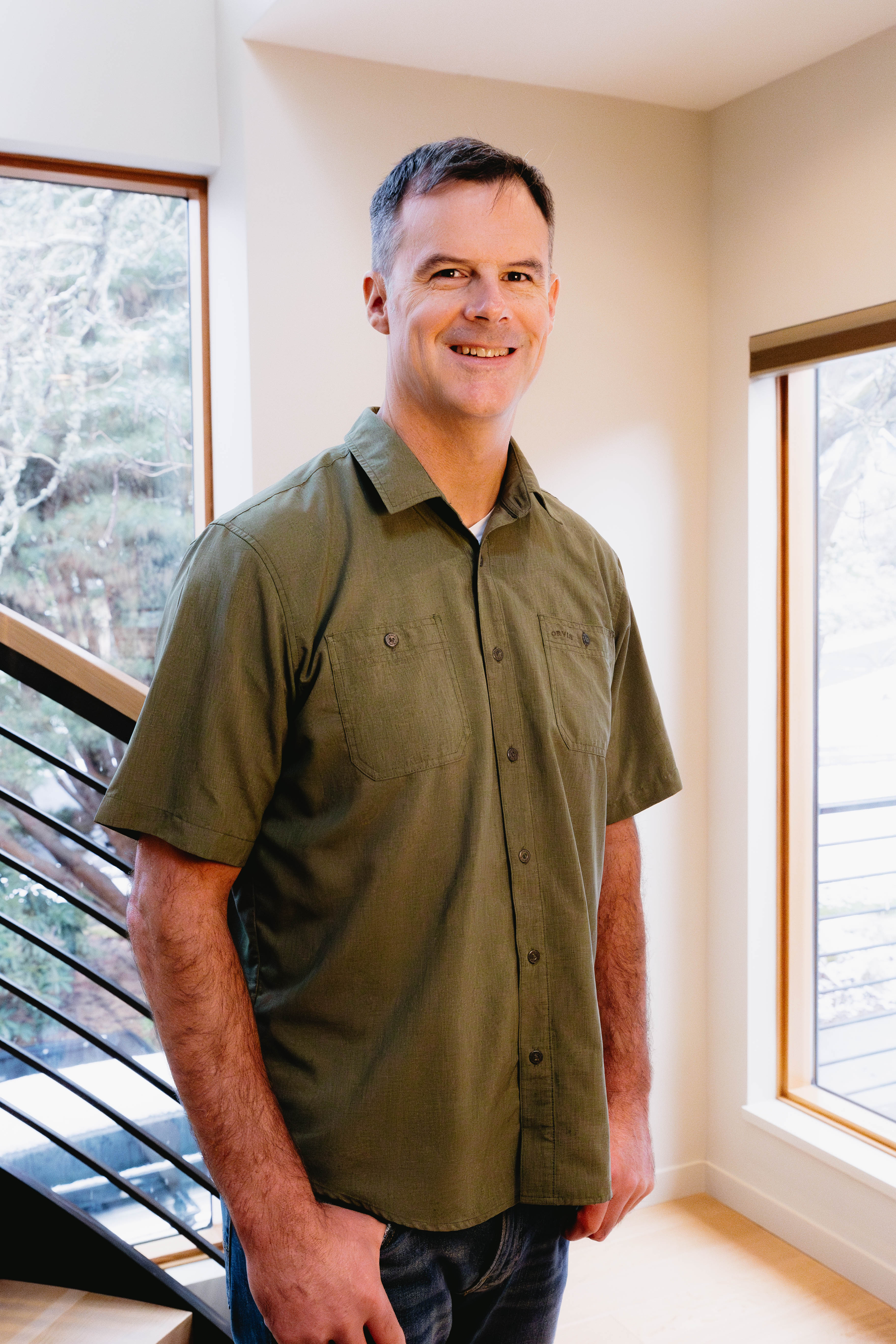 Man wearing a green shirt standing in a room with large windows.