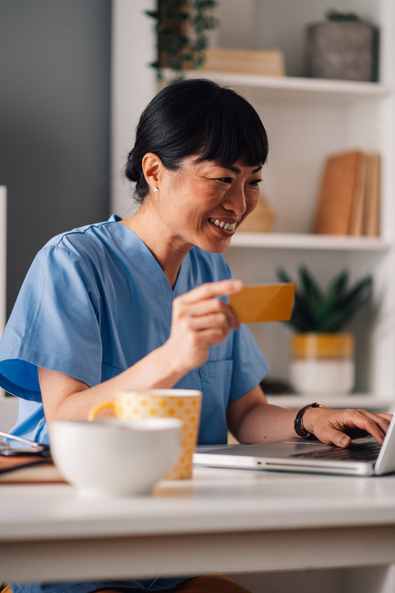 Person in blue scrubs holding a credit card and using a laptop at a desk.