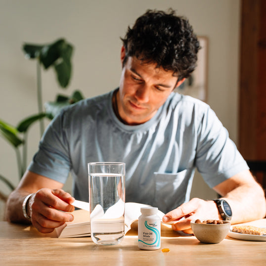 A man in a light blue t-shirt sits at a wooden table, looking down at a white bottle of dietary supplements alongside a glass of water, a small bowl, and an open book.