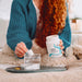 Person pouring Optima Collagen powder into a glass of water on a cozy couch.