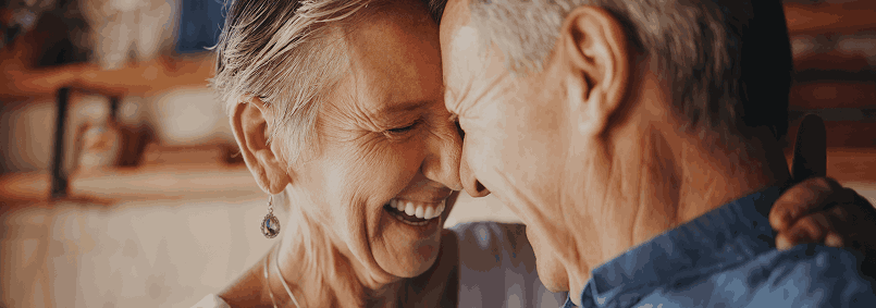Two elderly people smiling closely together, with a warm indoor setting.